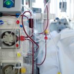 A close-up view of dialyzer tubes with blood flow and a blurry patient in the background, a sight that may be associated with Inadequate Dialysis.